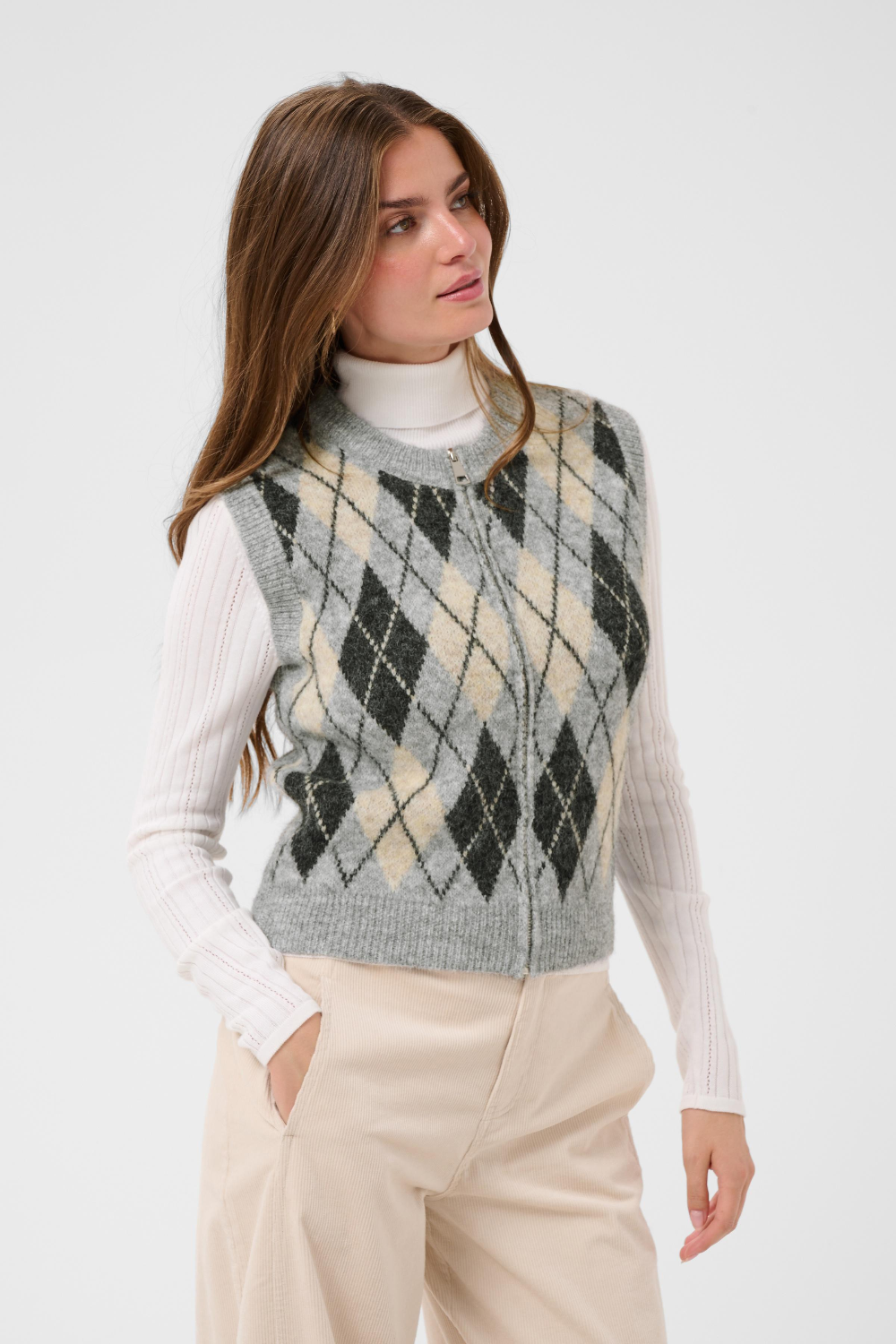 A woman wearing a gray zippered knit sweater vest with a beige and gray argyle pattern, layered over a white ribbed turtleneck and paired with beige trousers, against a plain white studio background.