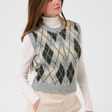 A woman wearing a gray zippered knit sweater vest with a beige and gray argyle pattern, layered over a white ribbed turtleneck and paired with beige trousers, against a plain white studio background.