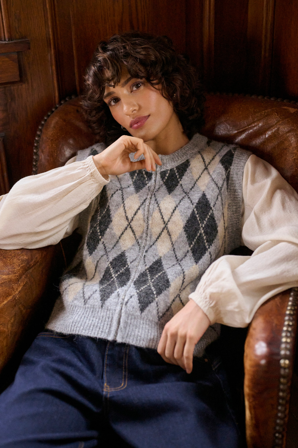 A person wearing a cream and grey argyle sweater vest over a cream blouse, paired with navy wide-leg trousers, sitting in a rustic wood-paneled room on a brown leather armchair.