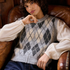 A person wearing a cream and grey argyle sweater vest over a cream blouse, paired with navy wide-leg trousers, sitting in a rustic wood-paneled room on a brown leather armchair.