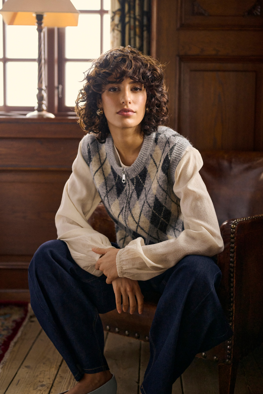A person wearing a cream and grey argyle sweater vest over a cream blouse, paired with navy wide-leg trousers and light gray shoes, sitting in a rustic wood-paneled room on a brown leather armchair.