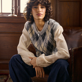 A person wearing a cream and grey argyle sweater vest over a cream blouse, paired with navy wide-leg trousers and light gray shoes, sitting in a rustic wood-paneled room on a brown leather armchair.