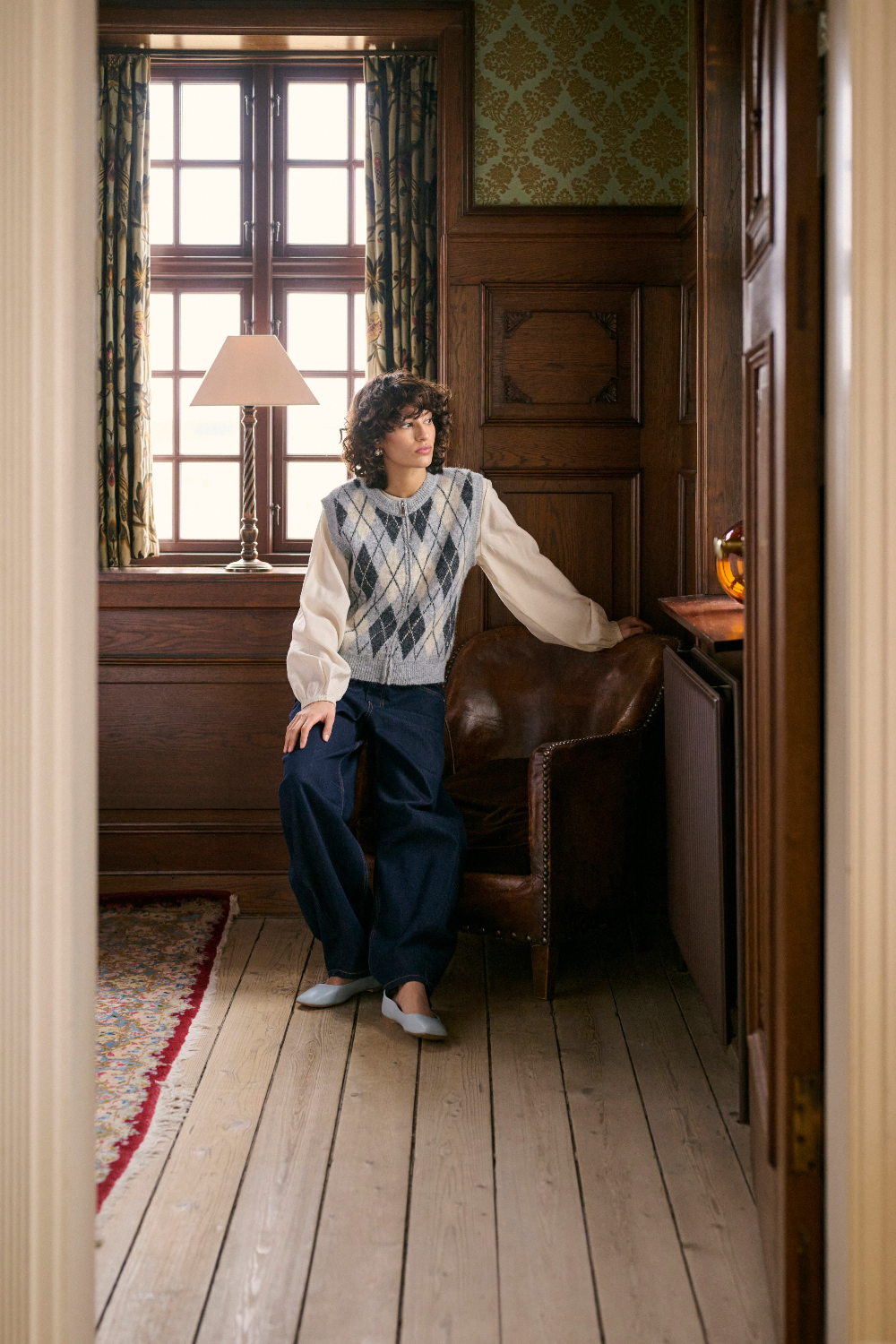 A person wearing a cream and grey argyle sweater vest over a cream blouse, paired with navy wide-leg trousers and light gray shoes, sitting in a rustic wood-paneled room next to a brown leather armchair.