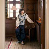 A person wearing a cream and grey argyle sweater vest over a cream blouse, paired with navy wide-leg trousers and light gray shoes, sitting in a rustic wood-paneled room next to a brown leather armchair.