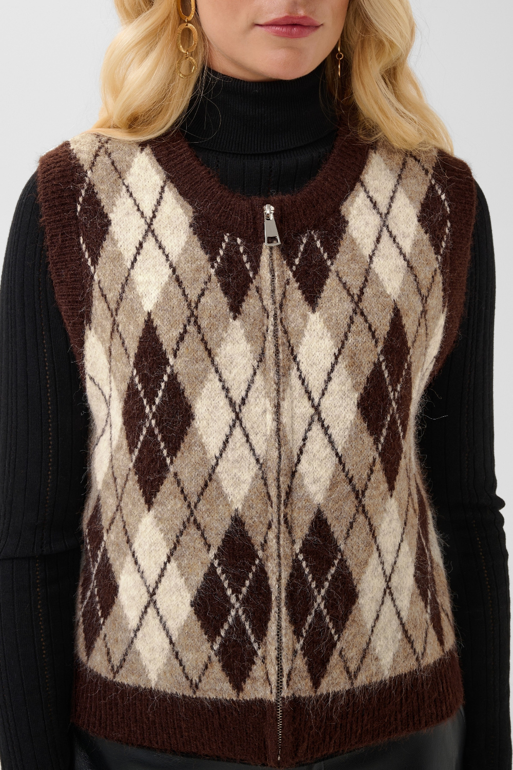 A close-up of a woman wearing a brown, beige, and cream argyle knit zip-up sweater vest over a black ribbed turtleneck.