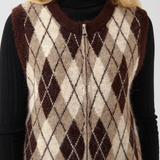A close-up of a woman wearing a brown, beige, and cream argyle knit zip-up sweater vest over a black ribbed turtleneck.