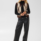 A woman modeling a brown and cream argyle knit sweater vest over a black ribbed turtleneck, paired with black faux leather pants and black boots, against a light gray studio background.