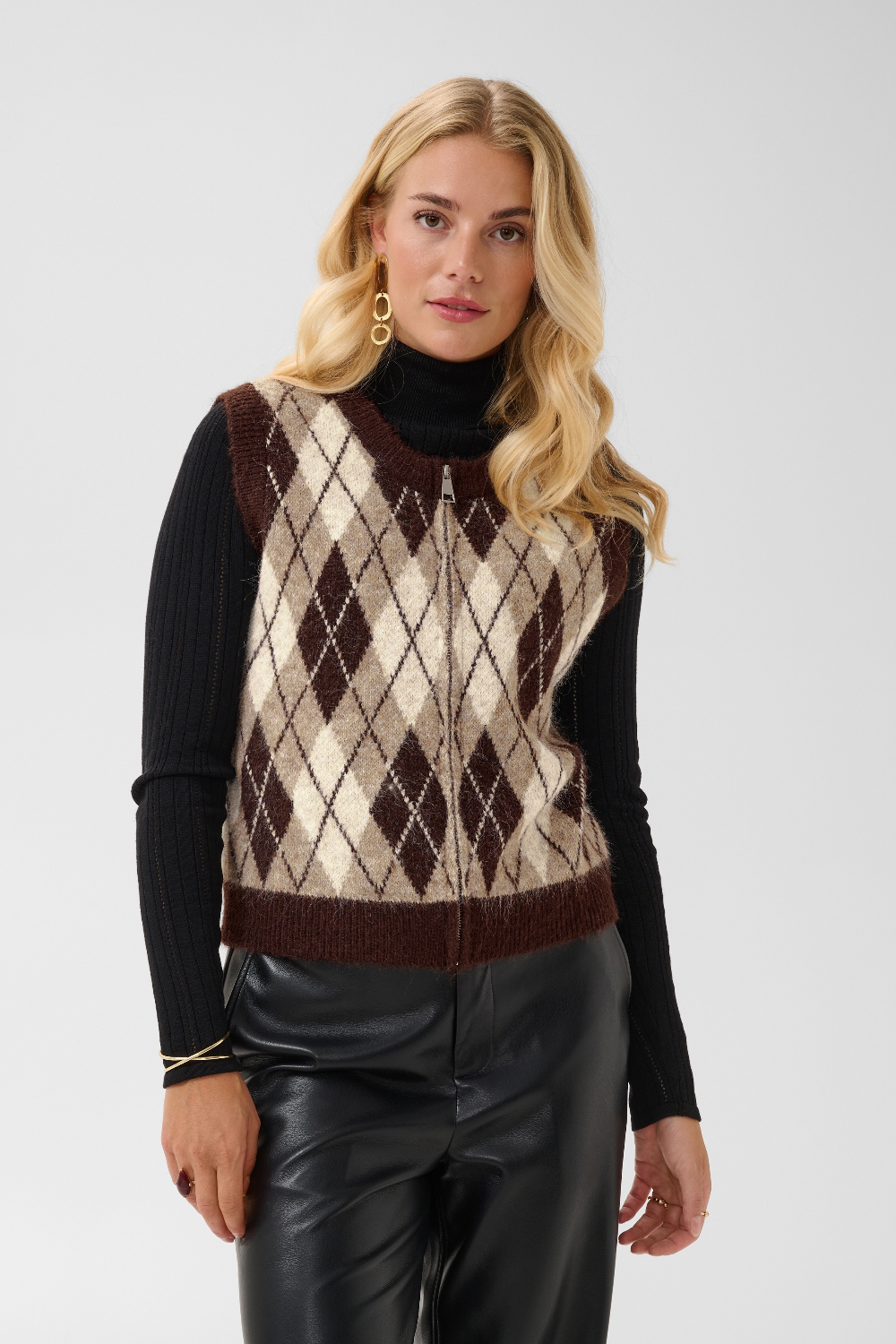 A woman modeling a brown and cream argyle knit sweater vest over a black ribbed turtleneck, paired with black faux leather pants, against a light gray studio background.