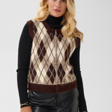 A woman modeling a brown and cream argyle knit sweater vest over a black ribbed turtleneck, paired with black faux leather pants, against a light gray studio background.