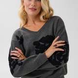 Woman modelling a grey v-neck sweater, with black accents, and black pants against a light background.
