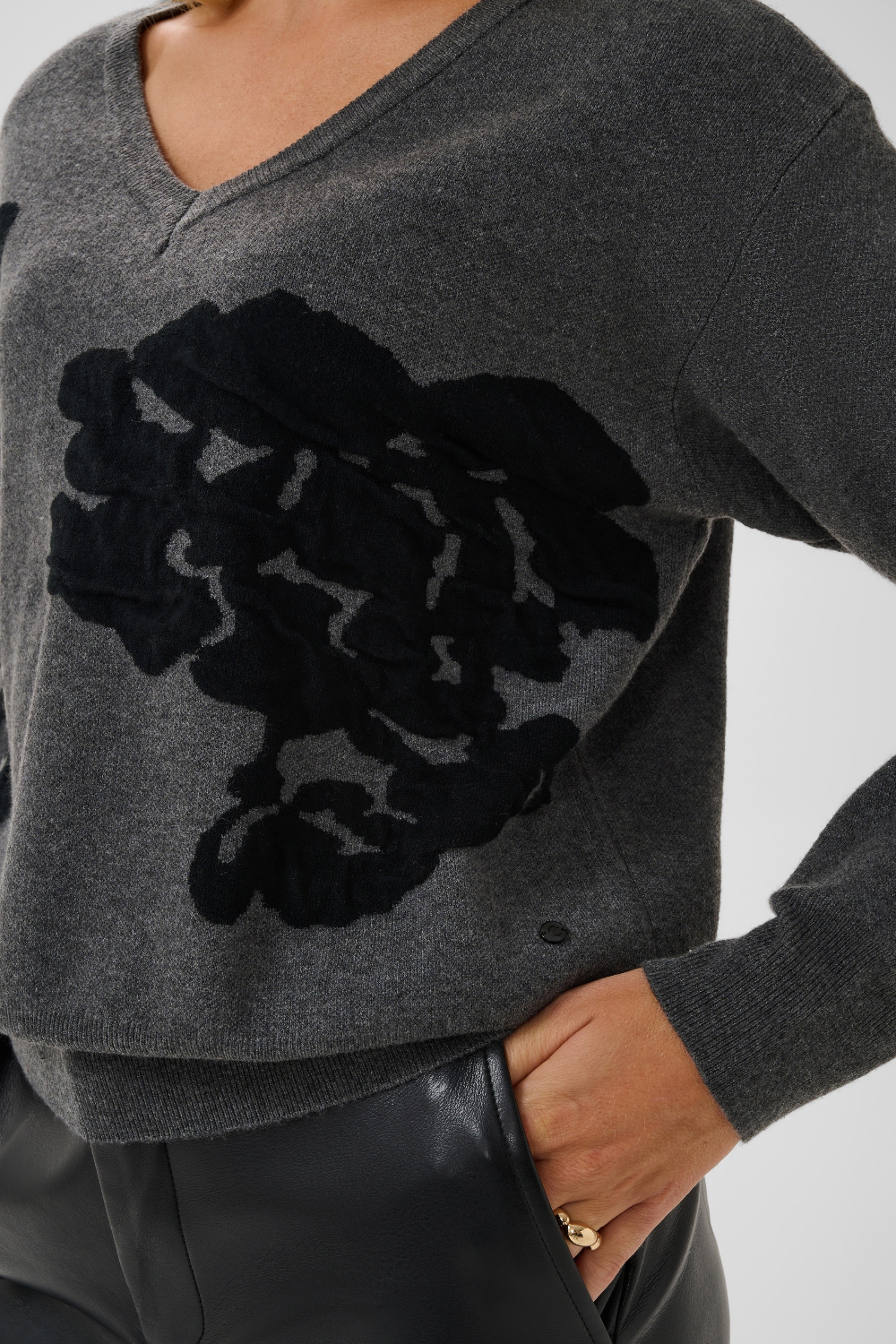 Close up of a woman modelling a grey v-neck sweater, with black accents, and black pants against a light background.