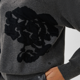 Close up of a woman modelling a grey v-neck sweater, with black accents, and black pants against a light background.
