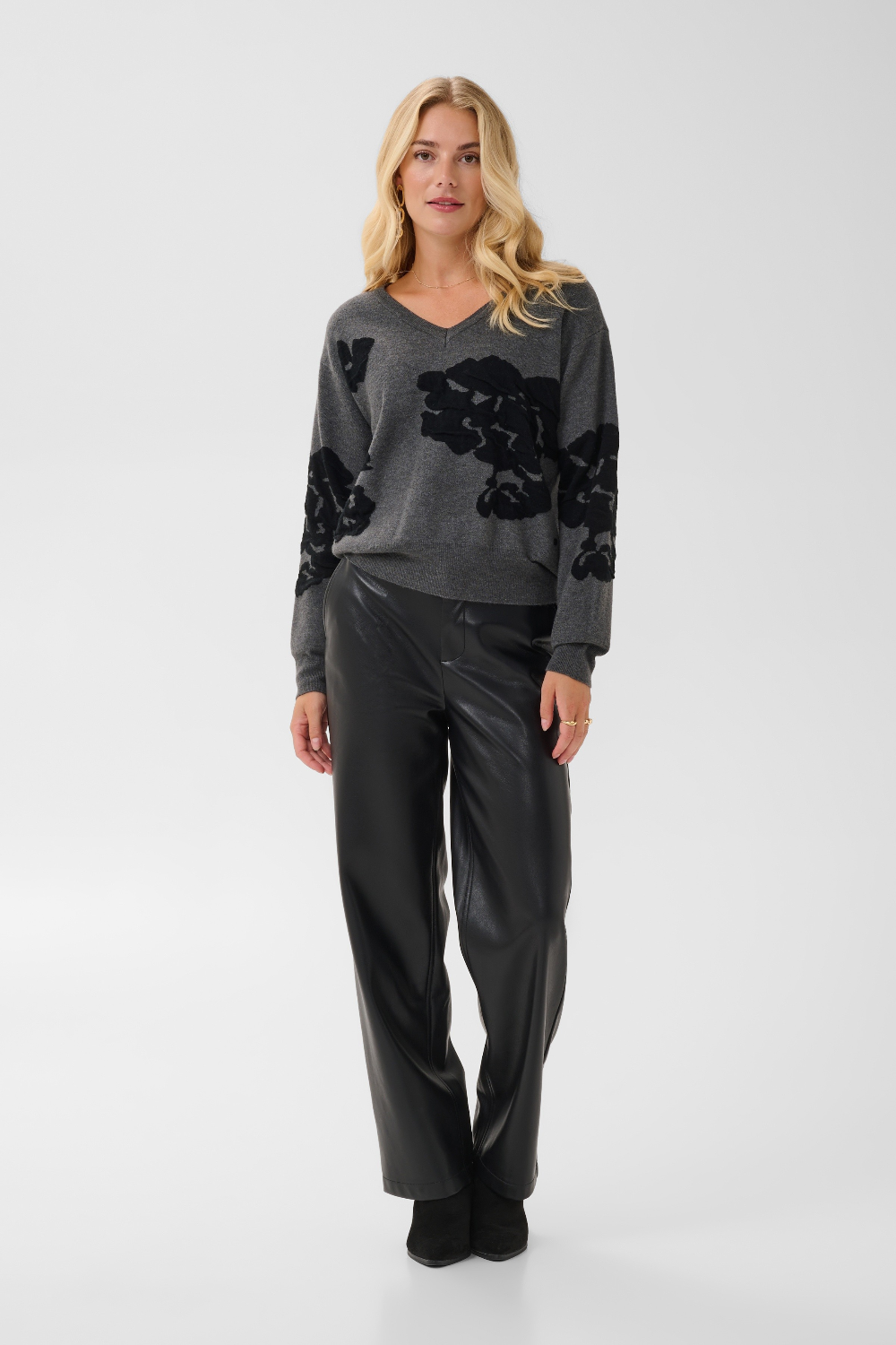 Woman modelling a grey v-neck sweater, with black accents, black pants and black boots against a light background.