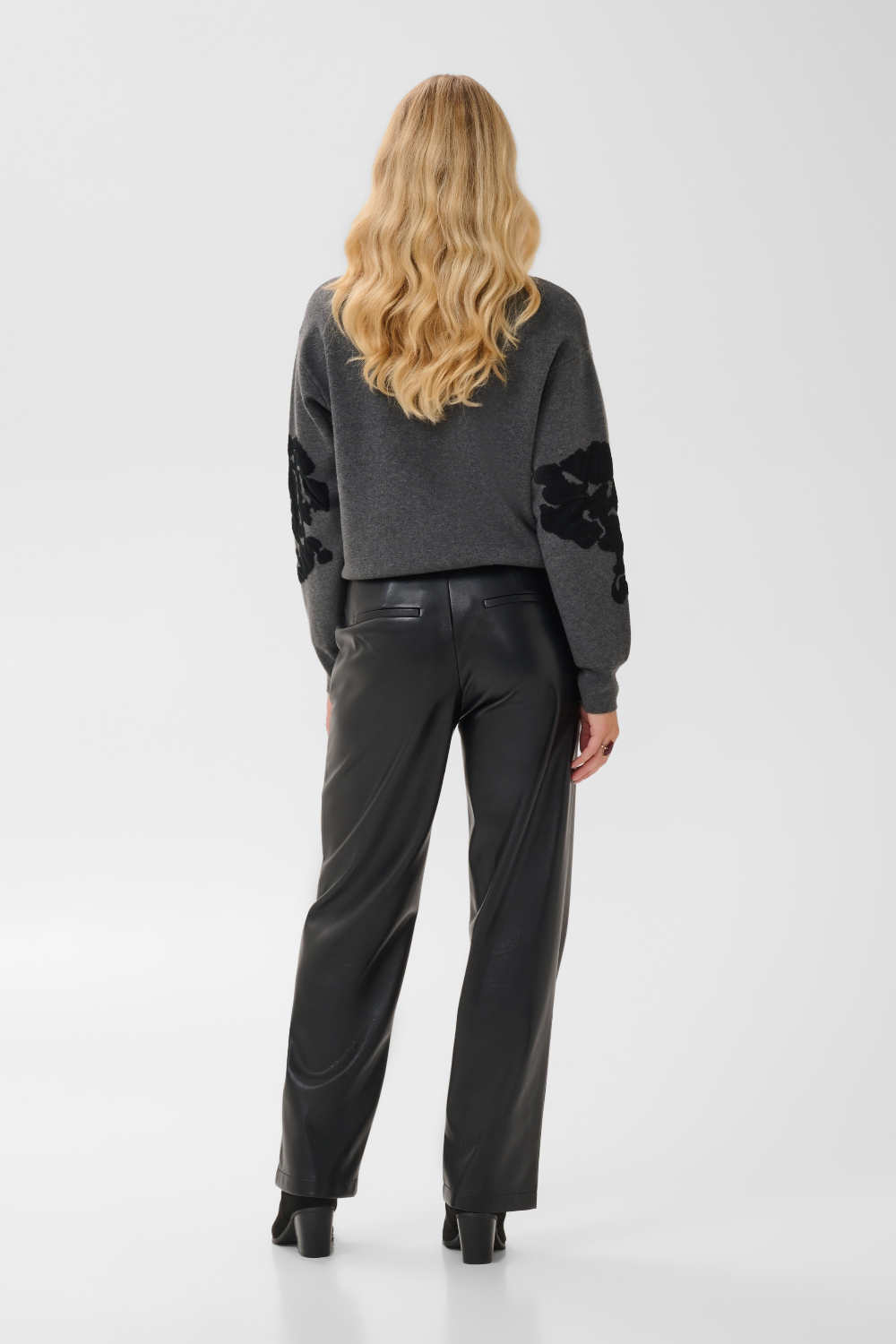 Woman, shown from behind, modelling a grey sweater, with black accents, black pants and black boots against a light background.