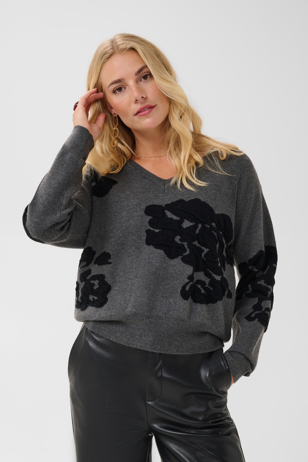 Woman modelling a grey v-neck sweater, with black accents, and black pants against a light background.