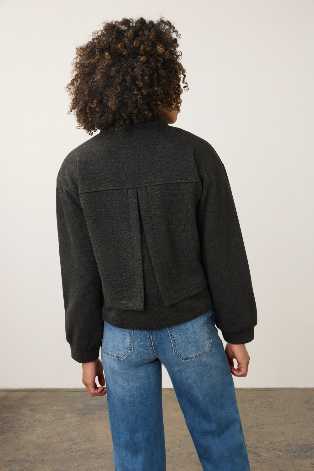 A person shown from the back wearing a dark charcoal gray cropped jacket with a unique vertical seam detail on the back, paired with blue jeans, against a plain off-white wall.