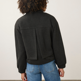 A person shown from the back wearing a dark charcoal gray cropped jacket with a unique vertical seam detail on the back, paired with blue jeans, against a plain off-white wall.