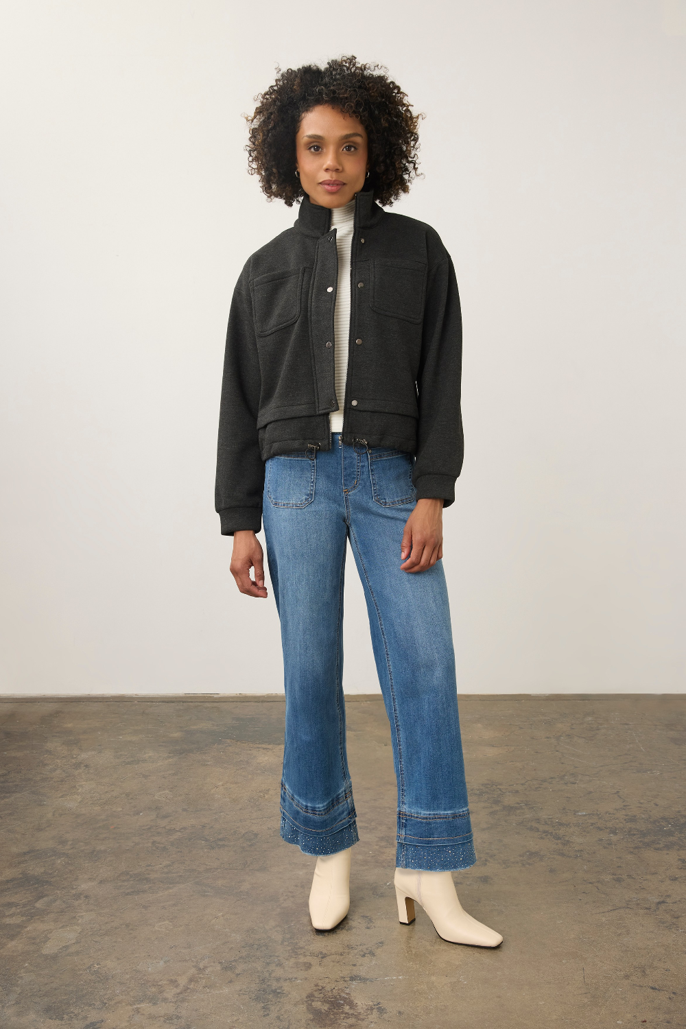 A model stands in a studio wearing a black cropped jacket over a white top, blue wide-leg jeans, and white heeled boots.