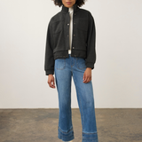 A model stands in a studio wearing a black cropped jacket over a white top, blue wide-leg jeans, and white heeled boots.