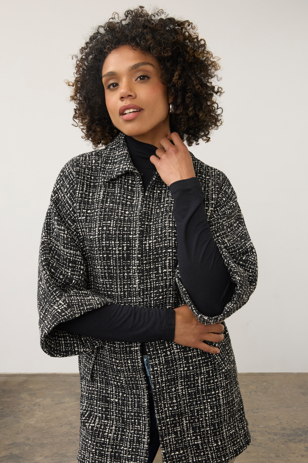 A woman with curly dark hair wearing a black and white textured tweed cape jacket over a black long-sleeve top, posing against a neutral light gray studio background.