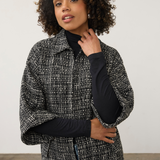 A woman with curly dark hair wearing a black and white textured tweed cape jacket over a black long-sleeve top, posing against a neutral light gray studio background.