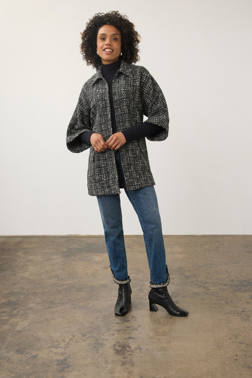 A woman modeling a black and gray textured tweed jacket with wide sleeves, worn over a black turtleneck, blue jeans, and black ankle boots, standing against a plain studio wall.