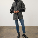 A woman modeling a black and gray textured tweed jacket with wide sleeves, worn over a black turtleneck, blue jeans, and black ankle boots, standing against a plain studio wall.