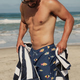 Man on a beach wearing navy swim trunks with fish pattern and holding a striped towel.