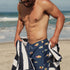Man on a beach wearing navy swim trunks with fish pattern and holding a striped towel.