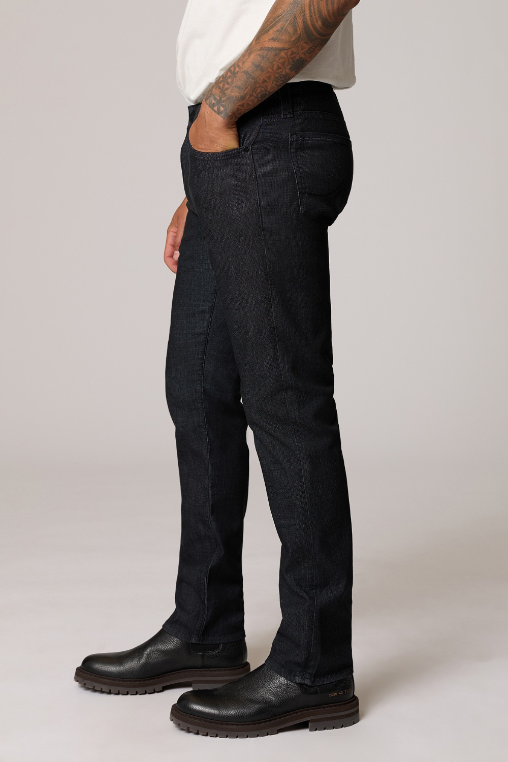 A side-profile view of a person wearing dark indigo slim-fit jeans, a white t-shirt, and black leather boots against a light gray studio background.