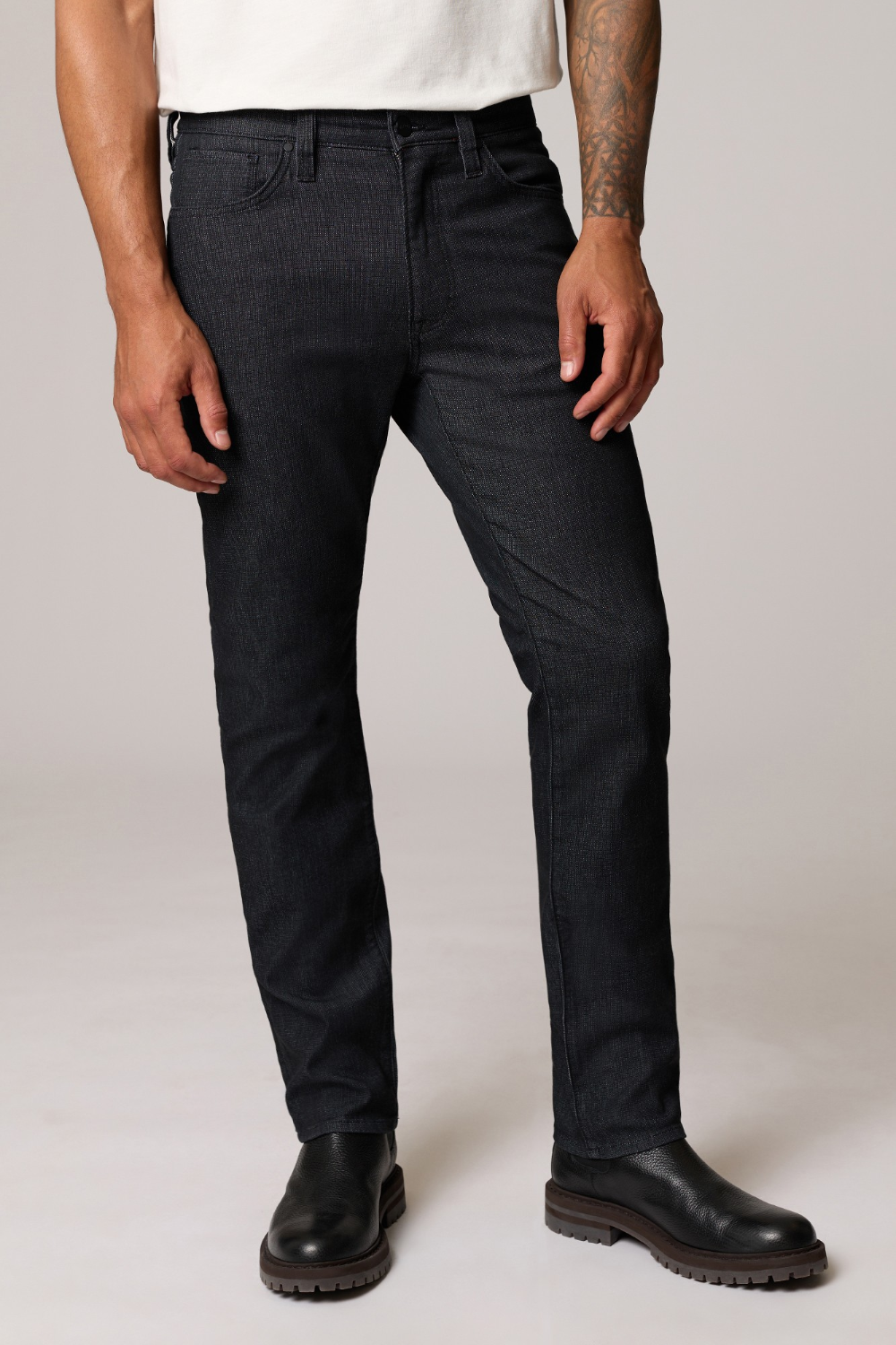 A man wearing slim-fit dark charcoal gray jeans, a white t-shirt, and black leather boots, standing against a light gray studio background.