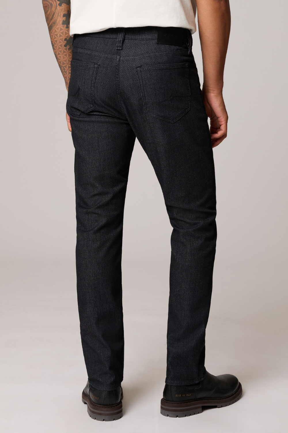 Rear view of a person wearing slim-fit dark indigo denim jeans, a white t-shirt, and black leather boots against a light gray studio background.