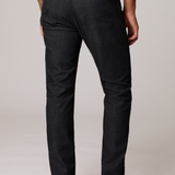 Rear view of a person wearing slim-fit dark indigo denim jeans, a white t-shirt, and black leather boots against a light gray studio background.