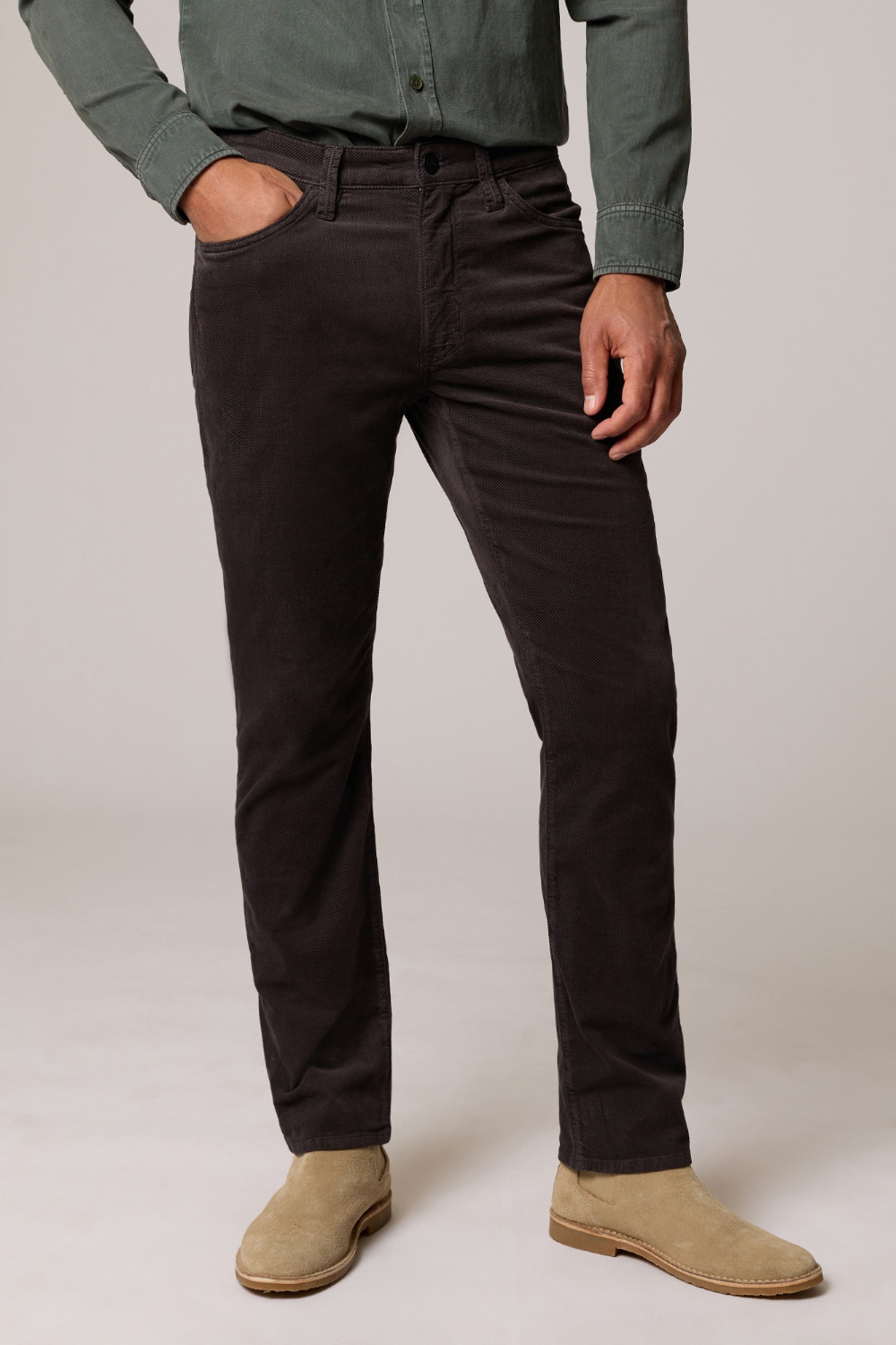A man wearing dark brown corduroy trousers, a sage green button-down shirt, and beige suede boots, standing against a light gray studio background.
