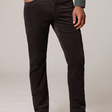 A man wearing dark brown corduroy trousers, a sage green button-down shirt, and beige suede boots, standing against a light gray studio background.