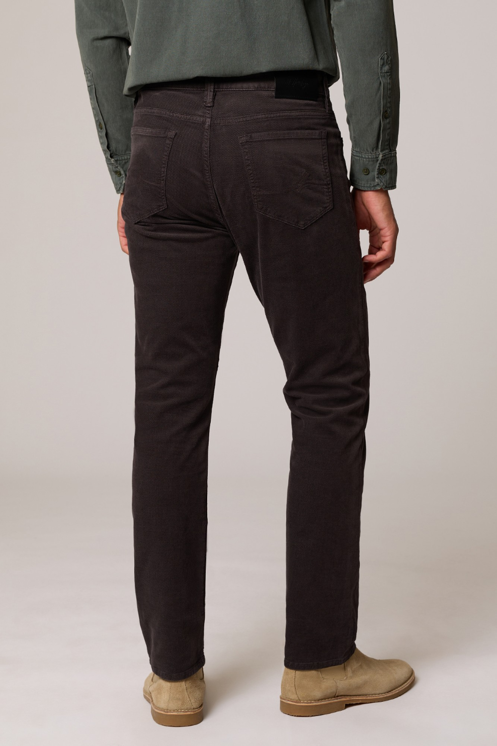 Rear view of a person wearing dark brown slim-fit jeans, an olive green shirt, and beige suede boots against a light gray studio background.