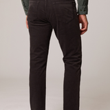 Rear view of a person wearing dark brown slim-fit jeans, an olive green shirt, and beige suede boots against a light gray studio background.