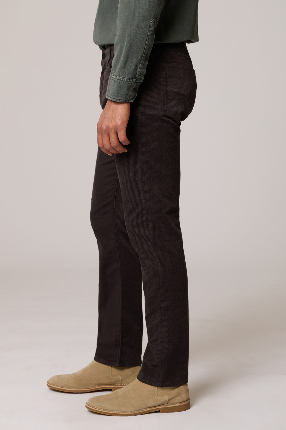 A side-profile view of a person wearing dark brown slim-fit corduroy trousers, a sage green shirt, and beige suede boots against a light gray studio background.