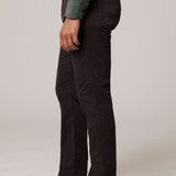 A side-profile view of a person wearing dark brown slim-fit corduroy trousers, a sage green shirt, and beige suede boots against a light gray studio background.