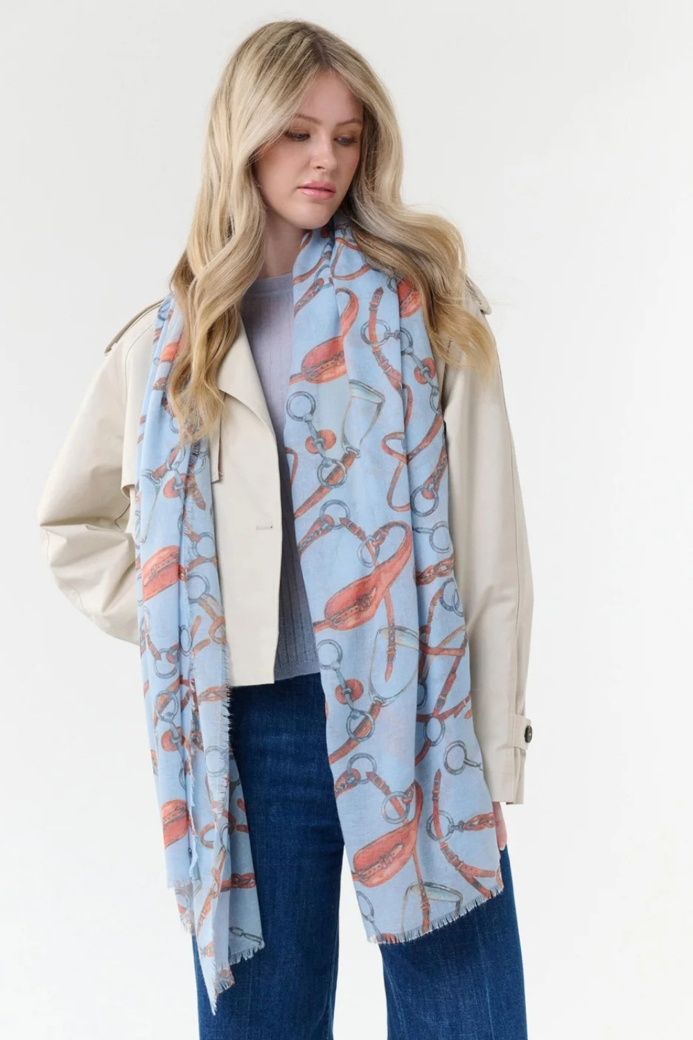 Woman modelling a blue coloured scarf with a brown and grey equestrian print, a blue top, blue jeans and a cream jacket, photographed on a light background.