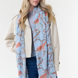 Woman modelling a blue coloured scarf with a brown and grey equestrian print, a blue top, blue jeans and a cream jacket, photographed on a light background.