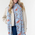 Woman modelling a blue coloured scarf with a brown and grey equestrian print, a blue top, blue jeans and a cream jacket, photographed on a light background.