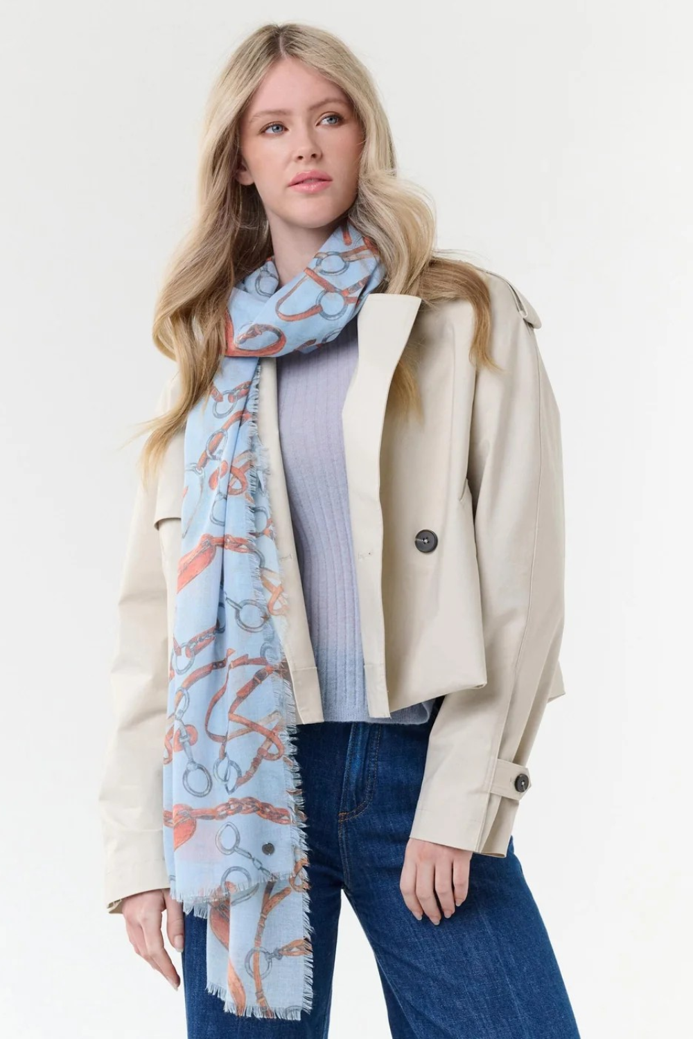 Woman modelling a blue coloured scarf with a brown and grey equestrian print, a blue top, blue jeans and a cream jacket, photographed on a light background.