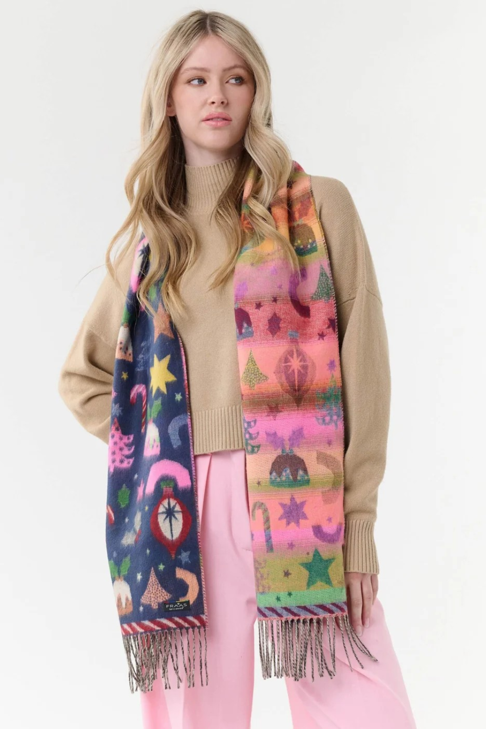 A woman modeling a long, multicolored Christmas-themed scarf with fringe, worn over a beige sweater and pink pants against a plain white studio background.