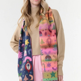 A woman modeling a long, multicolored Christmas-themed scarf with fringe, worn over a beige sweater and pink pants against a plain white studio background.