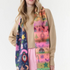 A woman modeling a long, multicolored Christmas-themed scarf with fringe, worn over a beige sweater and pink pants against a plain white studio background.