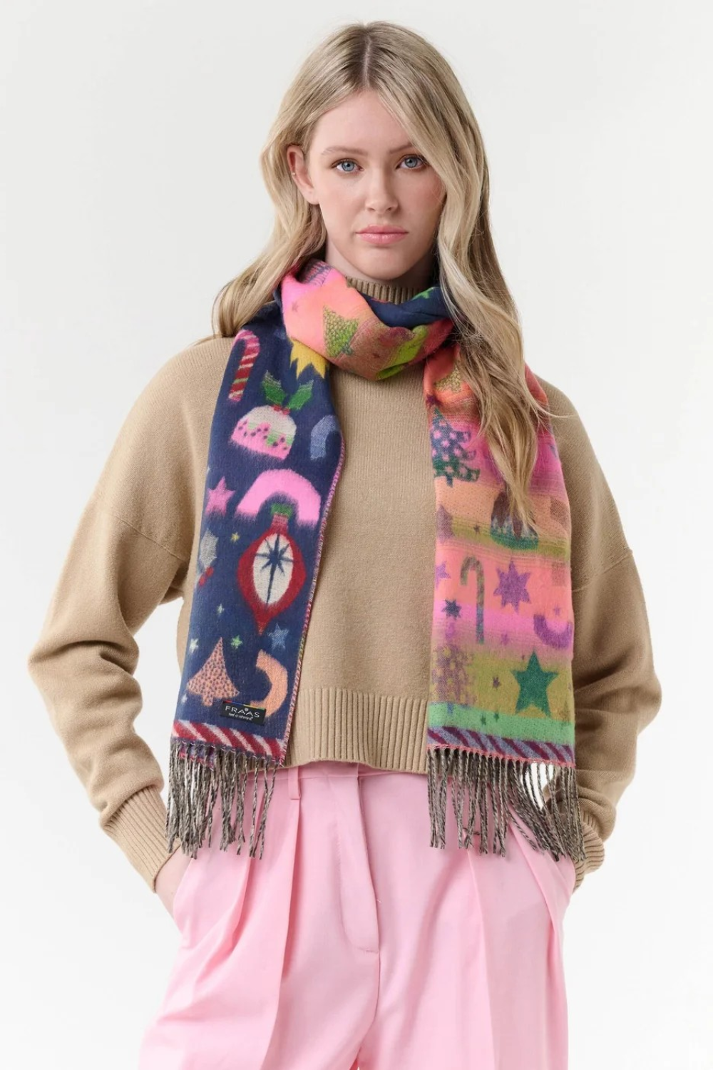 A woman modeling a vibrant, multicolored knit scarf with whimsical winter-themed patterns, including snowflakes, stars, and candy canes, styled over a beige sweater and pink trousers against a plain white studio background.