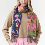 A woman modeling a vibrant, multicolored knit scarf with whimsical winter-themed patterns, including snowflakes, stars, and candy canes, styled over a beige sweater and pink trousers against a plain white studio background.