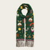 A dark green Christmas-themed scarf with a festive print of candy canes, stars, and mistletoe, finished with fringed ends, shown on a light beige background.
