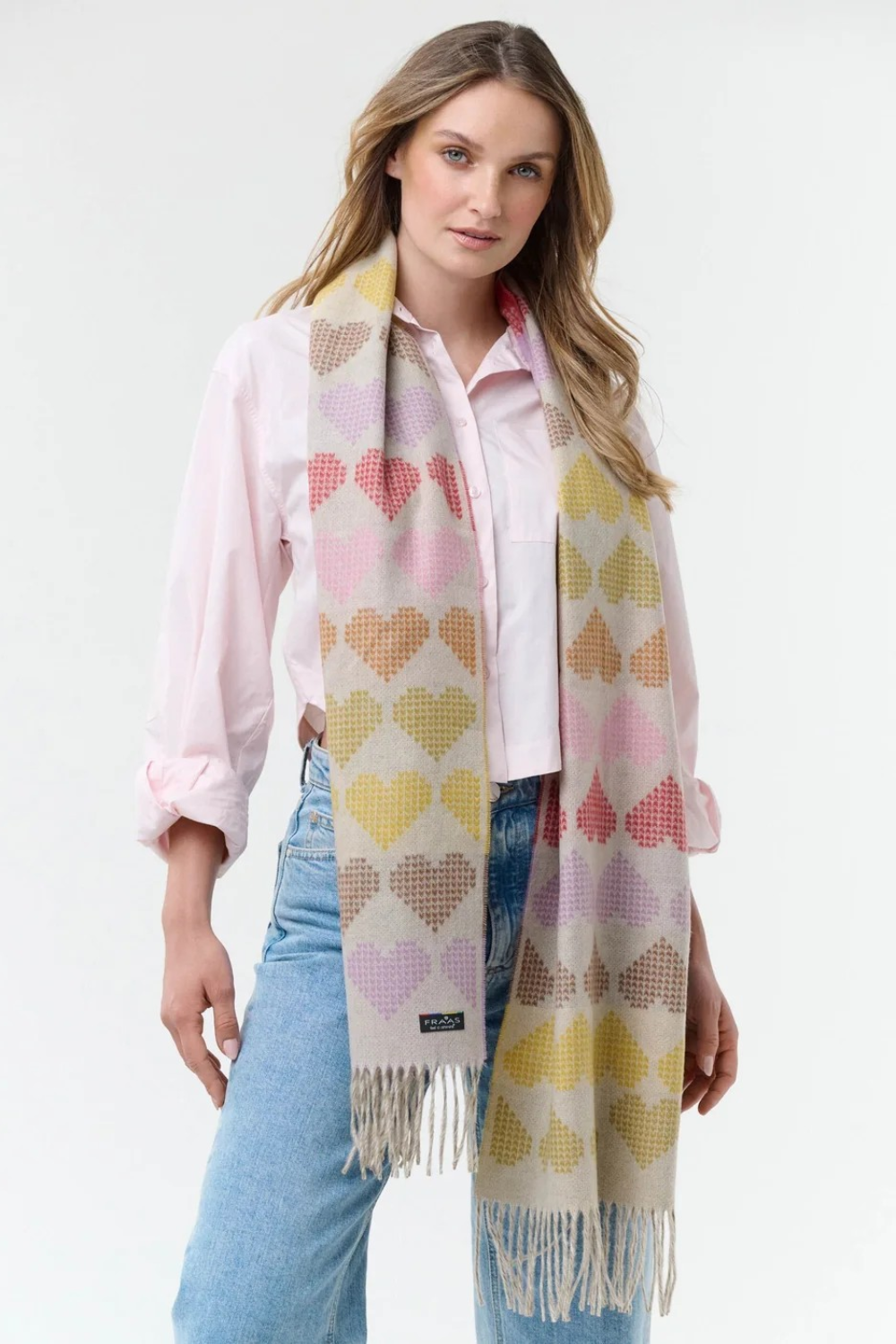 Woman modelling a multi coloured heart printed scarf, with fringe, with a pink blouse and blue jeans photographed on a light background.